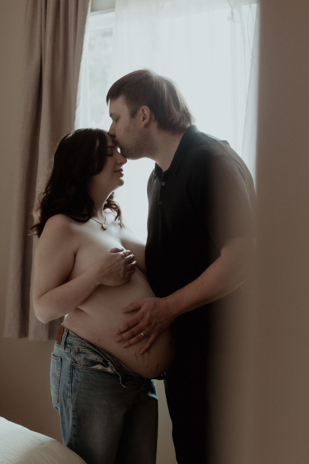 An intimate maternity portrait of an expecting couple sharing a tender moment by a window. PNW Maternity Photographer