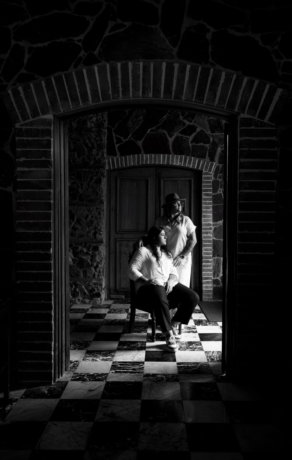 Artistic black and white portrait showing a person seated under a brick archway on checkered floor.