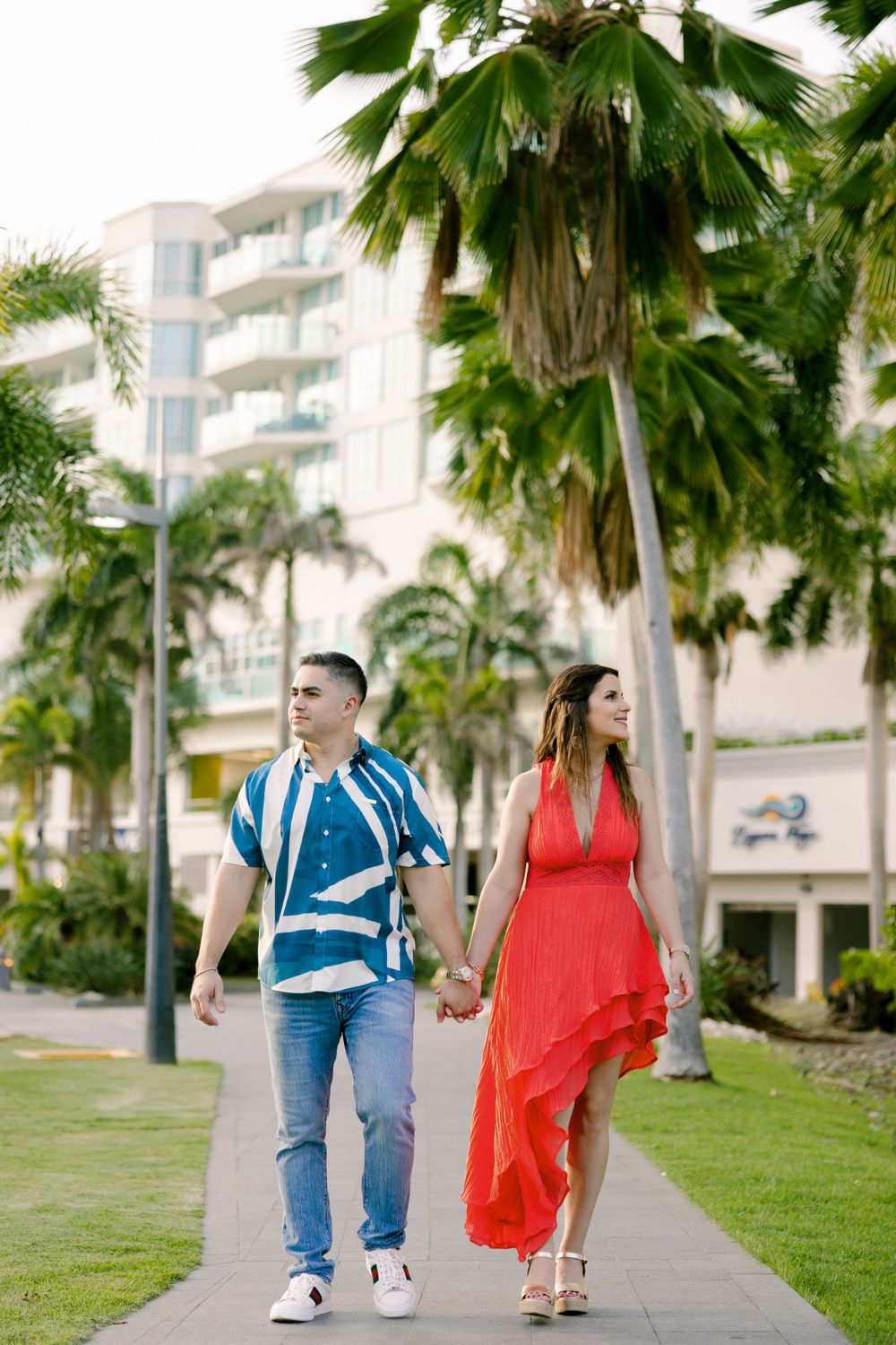 A couple walking hand in hand along a palm-lined path by luxury apartments.