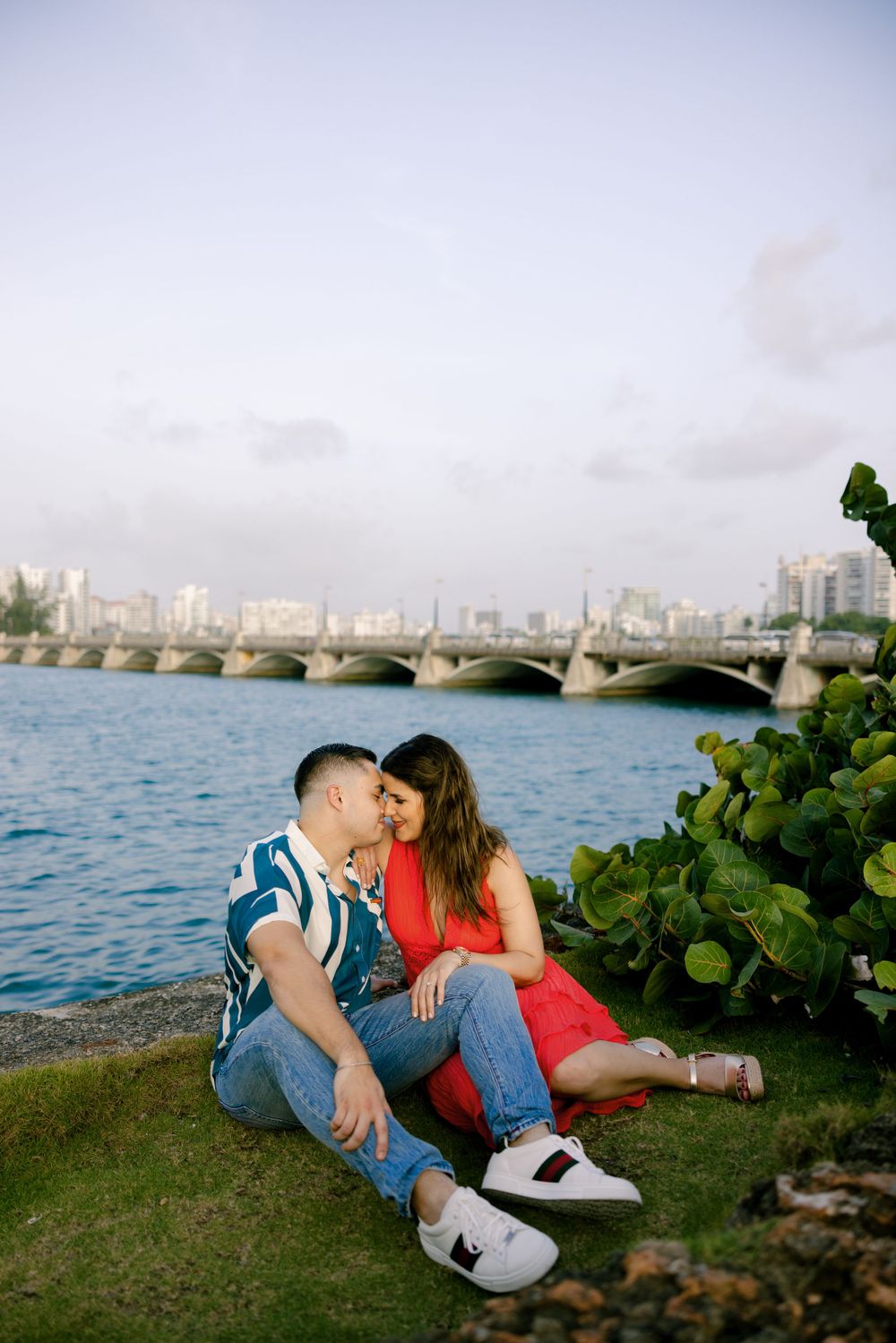 A couple sharing an intimate moment by the water with a city bridge in the background.