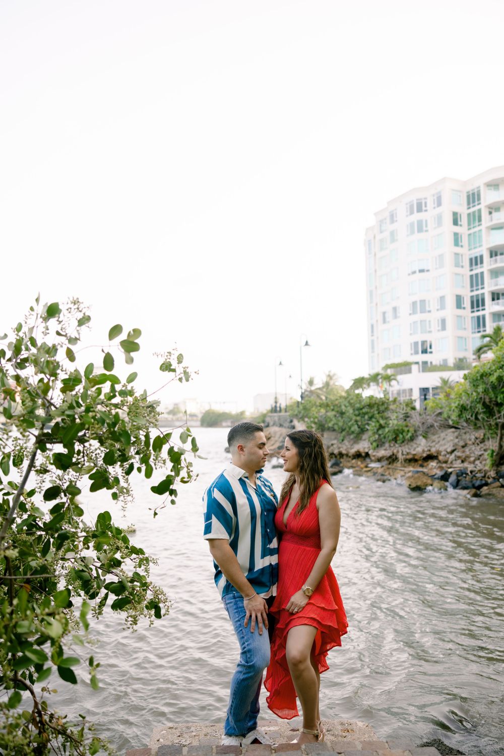A couple stands by the water's edge with a red dress flowing in the breeze near high-rise buildings.