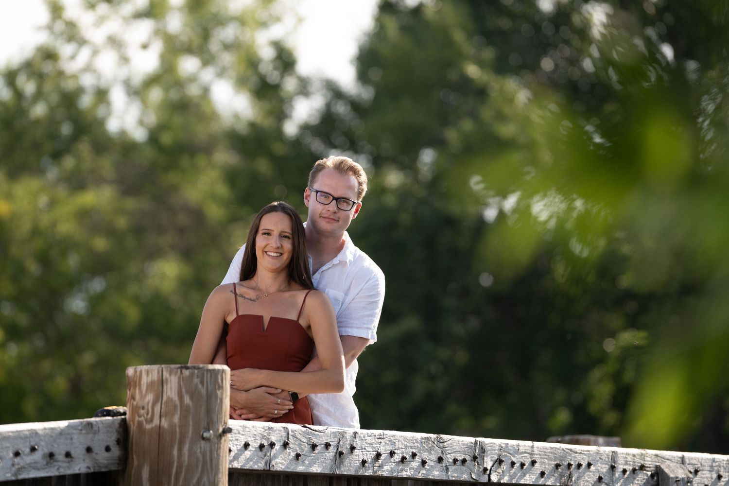 A couple sits together on a rustic wooden fence during a sunny outdoor photo session.