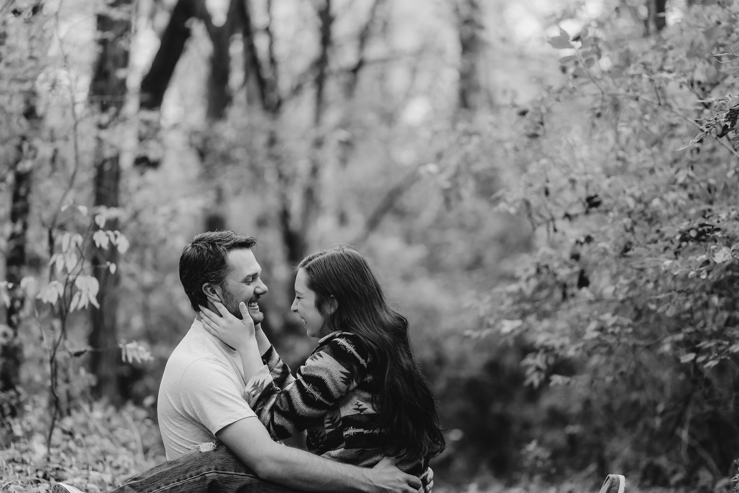 A joyful couple shares a laugh while embracing in a wooded setting during their black and white photoshoot.