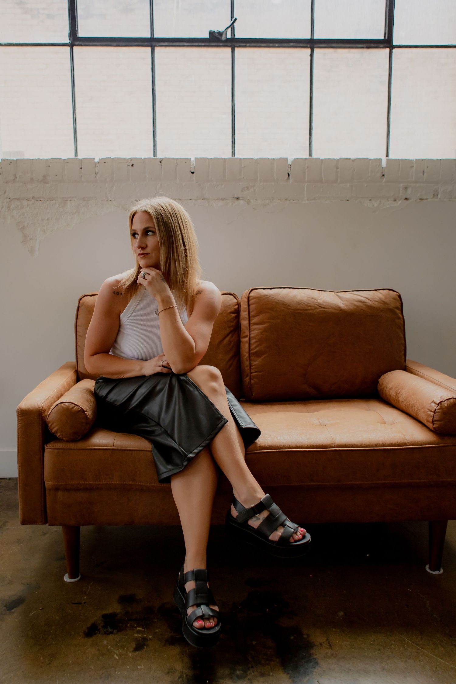 Woman in white top and dark skirt lounges on brown leather couch in industrial space.