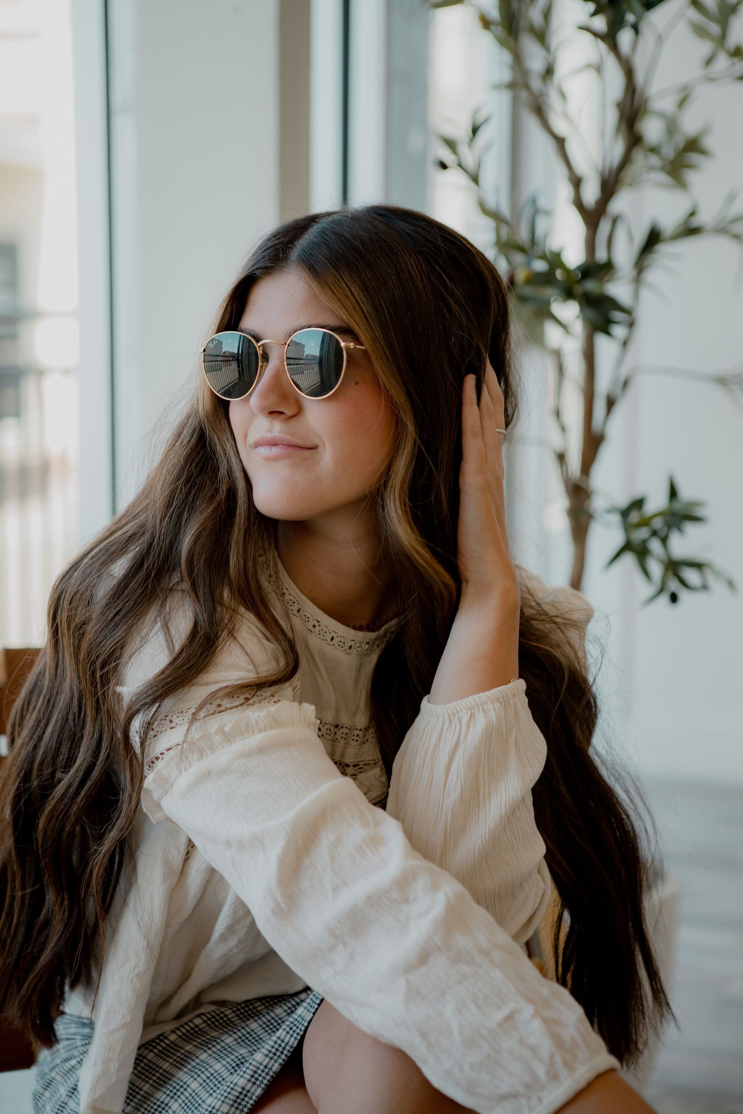 Woman with long dark hair wearing round sunglasses and white blouse sits by window with indoor plants.