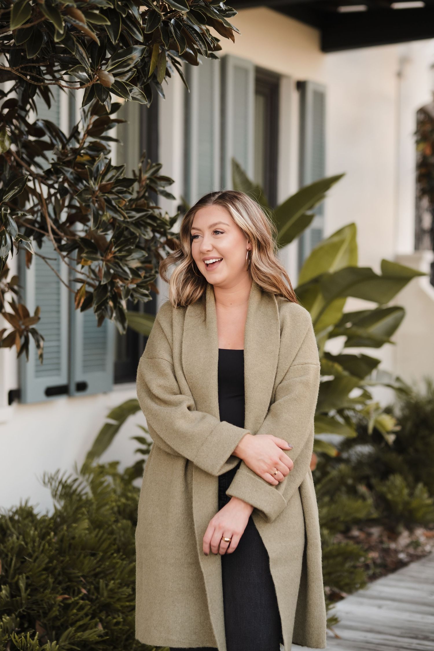 Woman in beige blazer laughing while posing against exterior of building with magnolia trees.