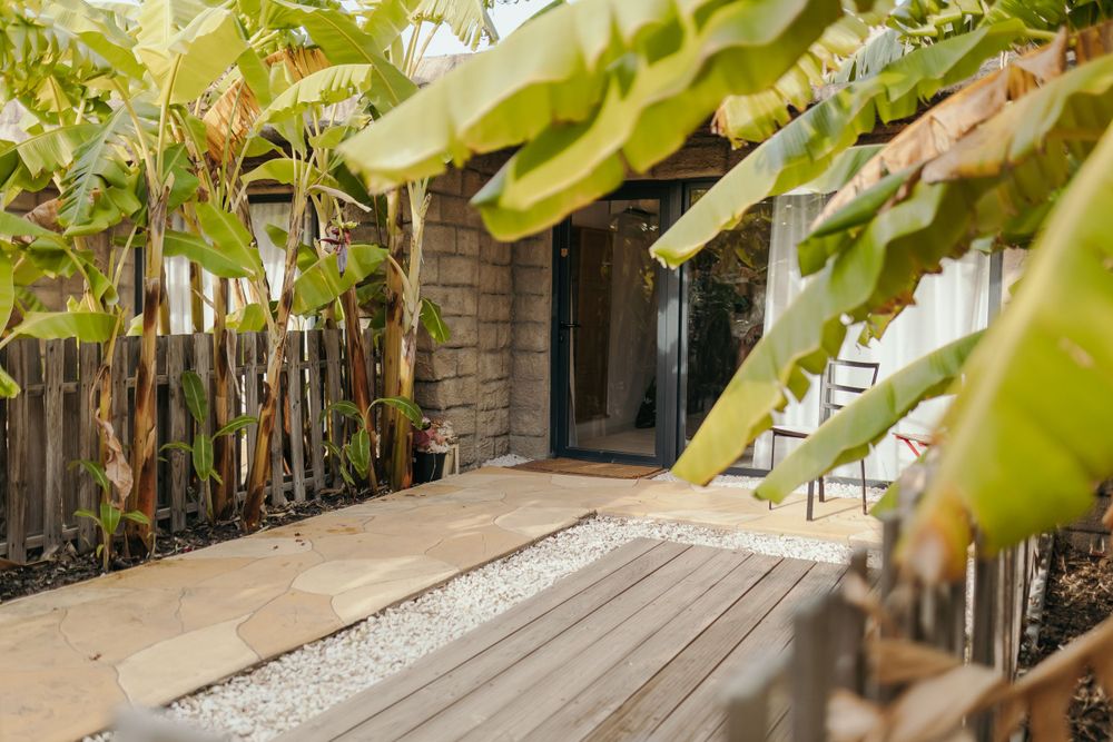 Tropical garden pathway with banana leaves framing a wooden deck leading to a cottage door.