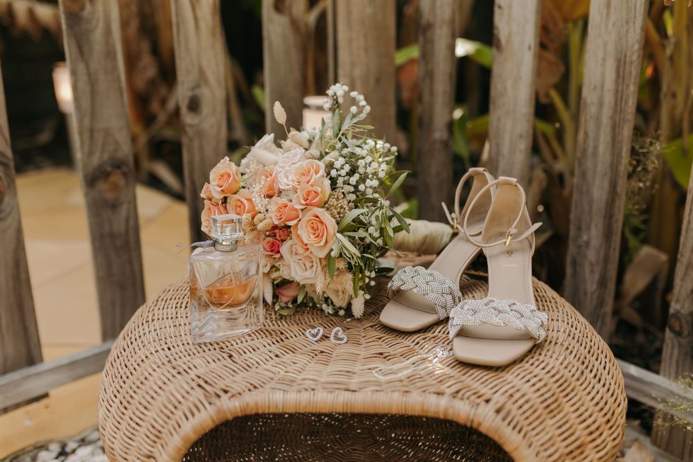 Bridal accessories including peach rose bouquet, white heels, and rings on a wicker table.