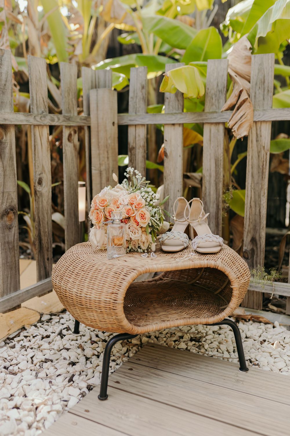 Bridal bouquet of peach roses and white heels displayed on a wicker table against a wooden fence.
