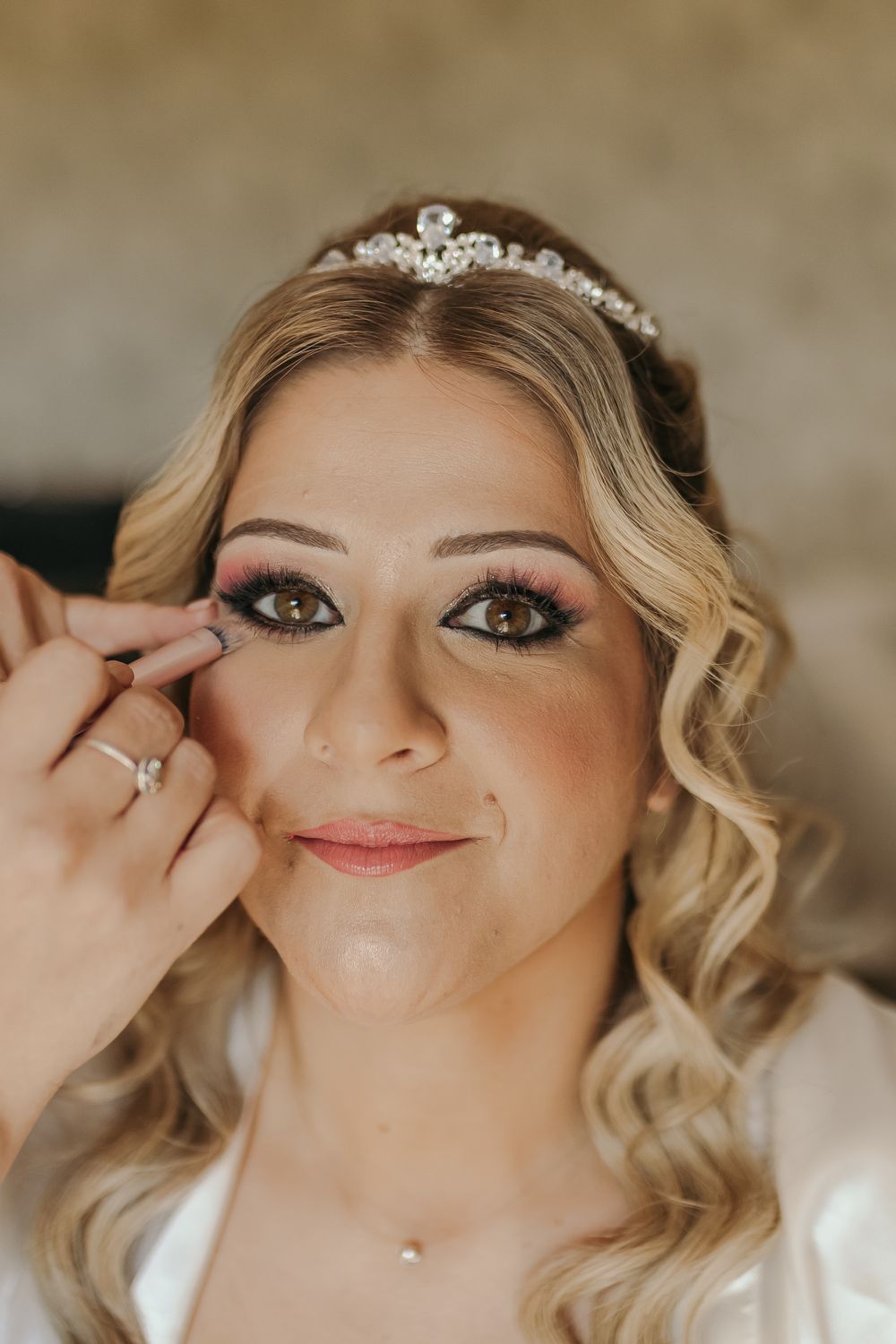 Close-up of a bride wearing a crystal tiara getting false eyelashes applied by a makeup artist.