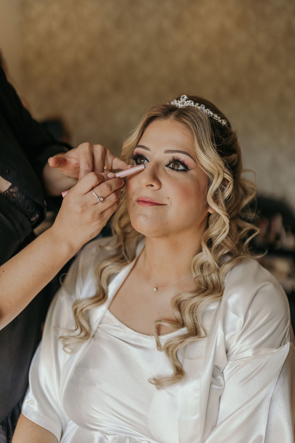 Bride in white robe wearing a crystal tiara getting eye makeup applied during bridal preparations.