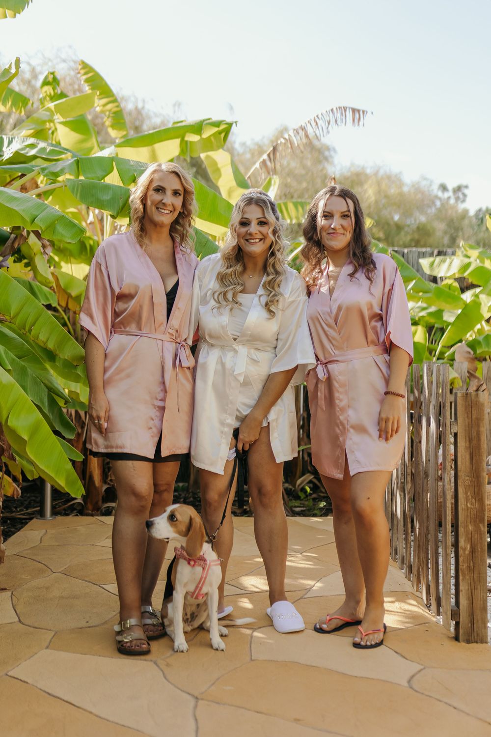 Bride in white robe poses with two bridesmaids in pink robes and a beagle dog on a garden pathway.