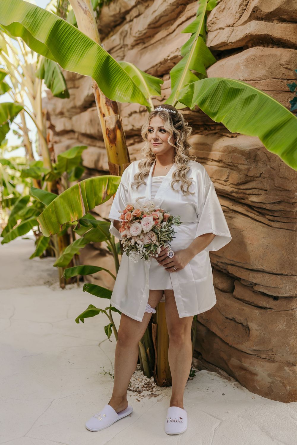 Smiling bride in white satin robe holds a pink rose bouquet beside tropical banana plants and stone wall.