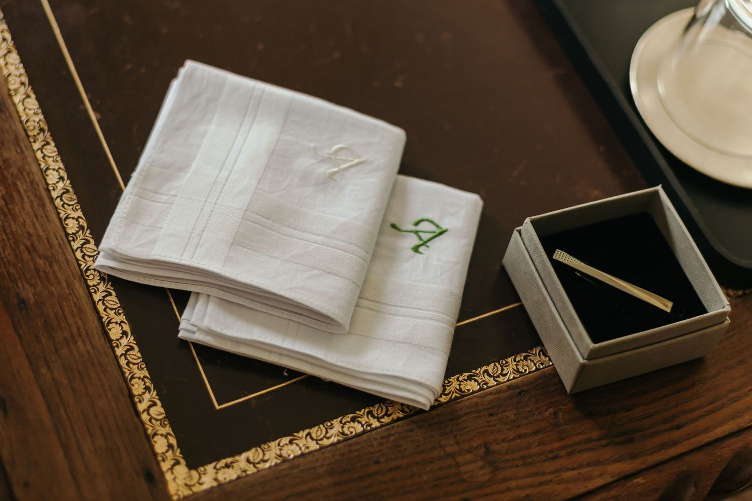 Elegant wedding details including monogrammed white handkerchiefs and gold tie clip in gift box on wooden desk.