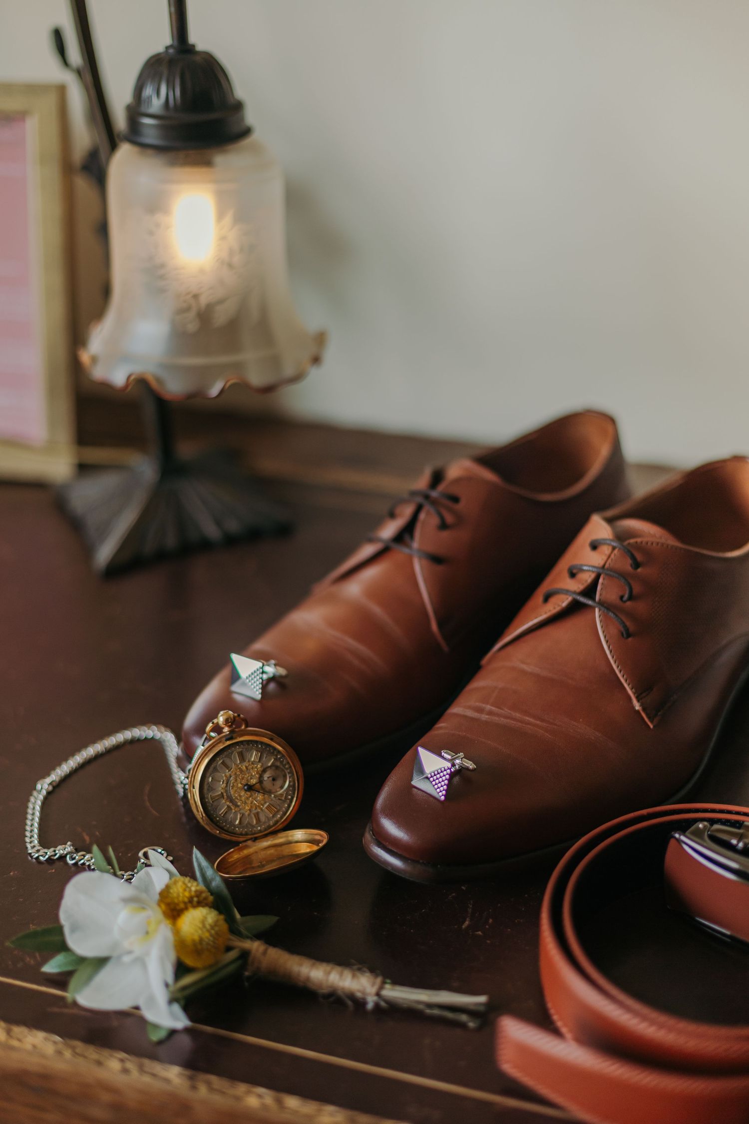 Brown leather dress shoes with cufflinks, pocket watch, boutonniere, and belt on wooden table.