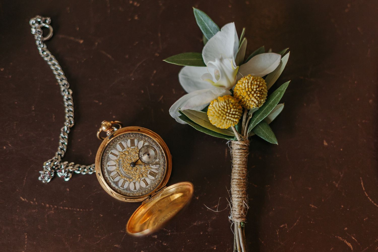 Gold vintage pocket watch with chain beside a boutonniere with white flowers and yellow billy buttons.