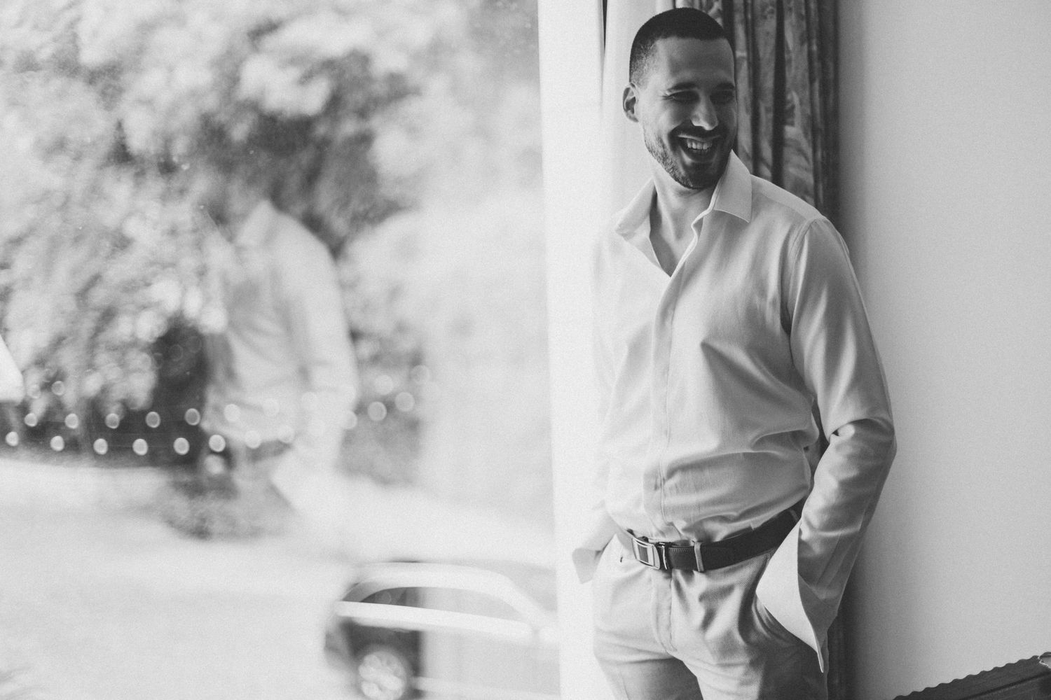 Smiling groom in white shirt leaning against a column in black and white wedding photo.