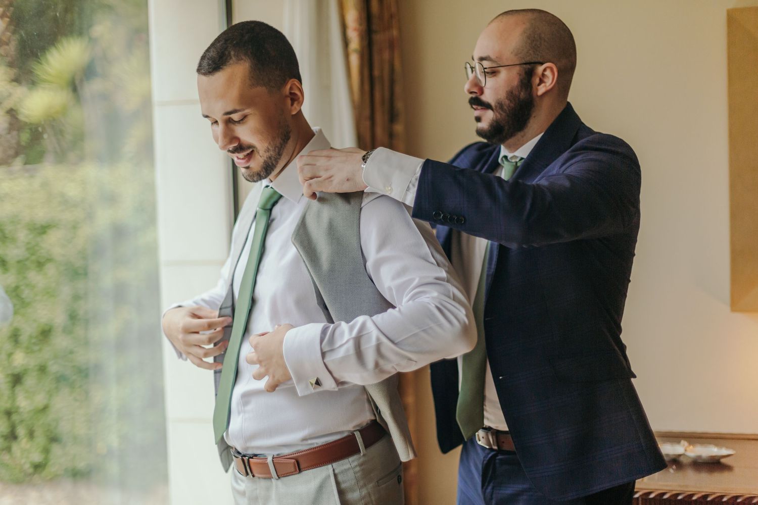Groomsman in navy suit helping groom in grey vest and green tie with his jacket.