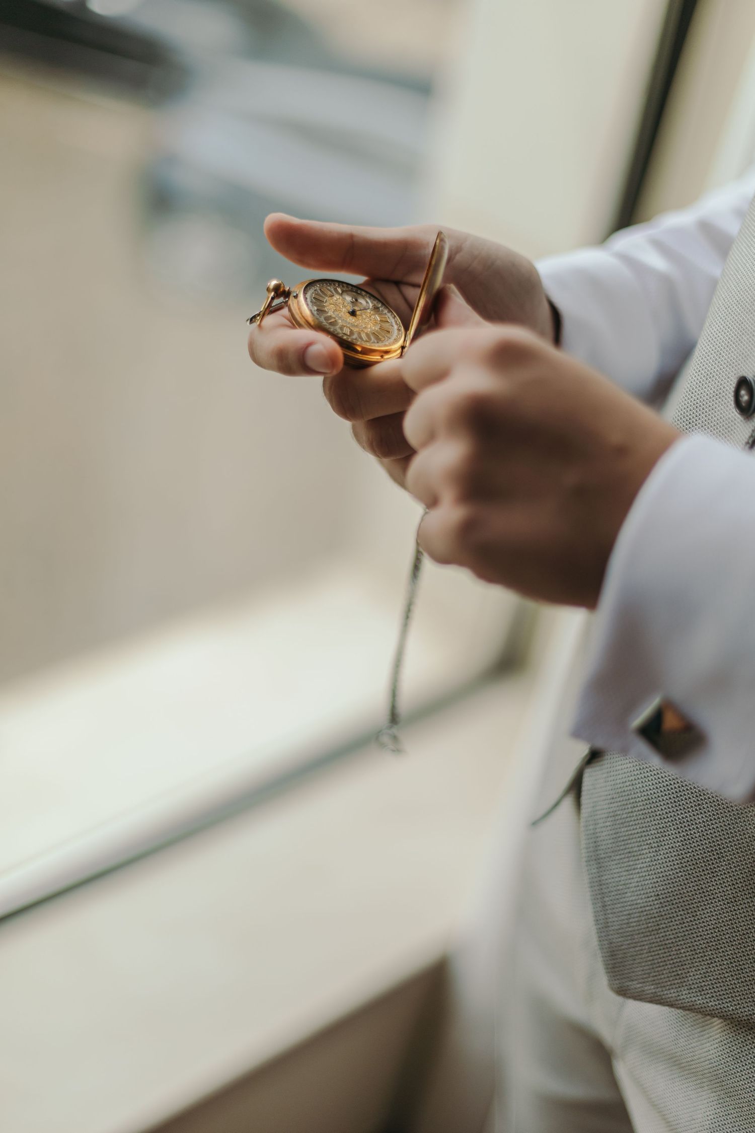 Groom in grey vest holding open gold vintage pocket watch near bright window.