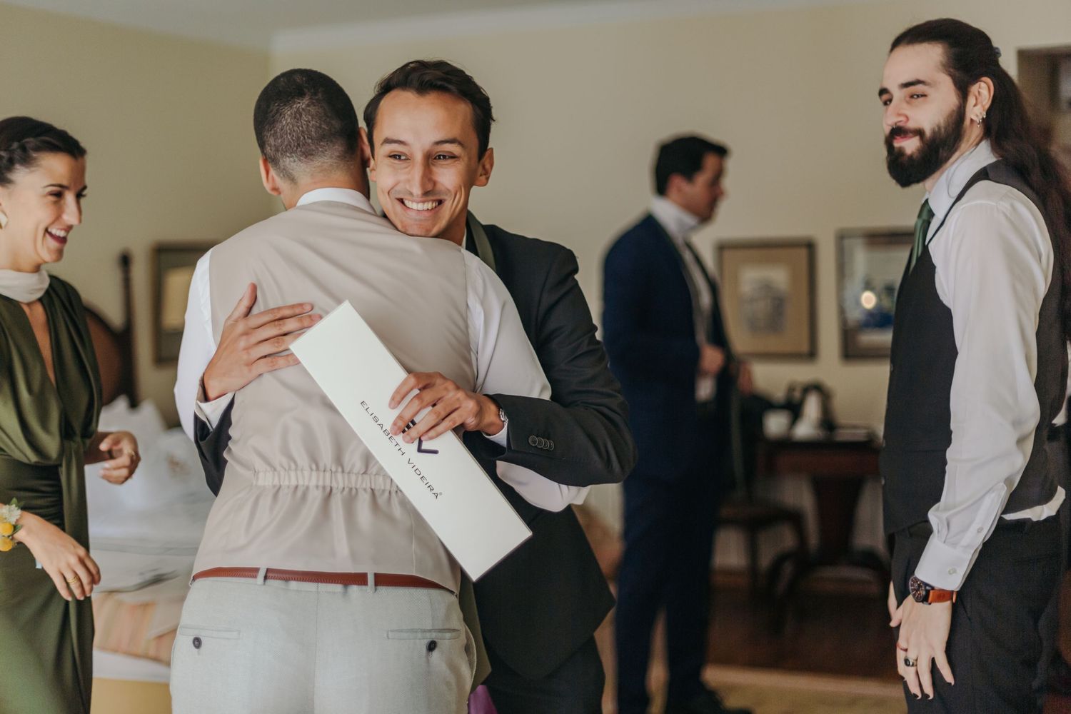 Groom in grey vest hugging a guest while others socialize in wedding preparation room.