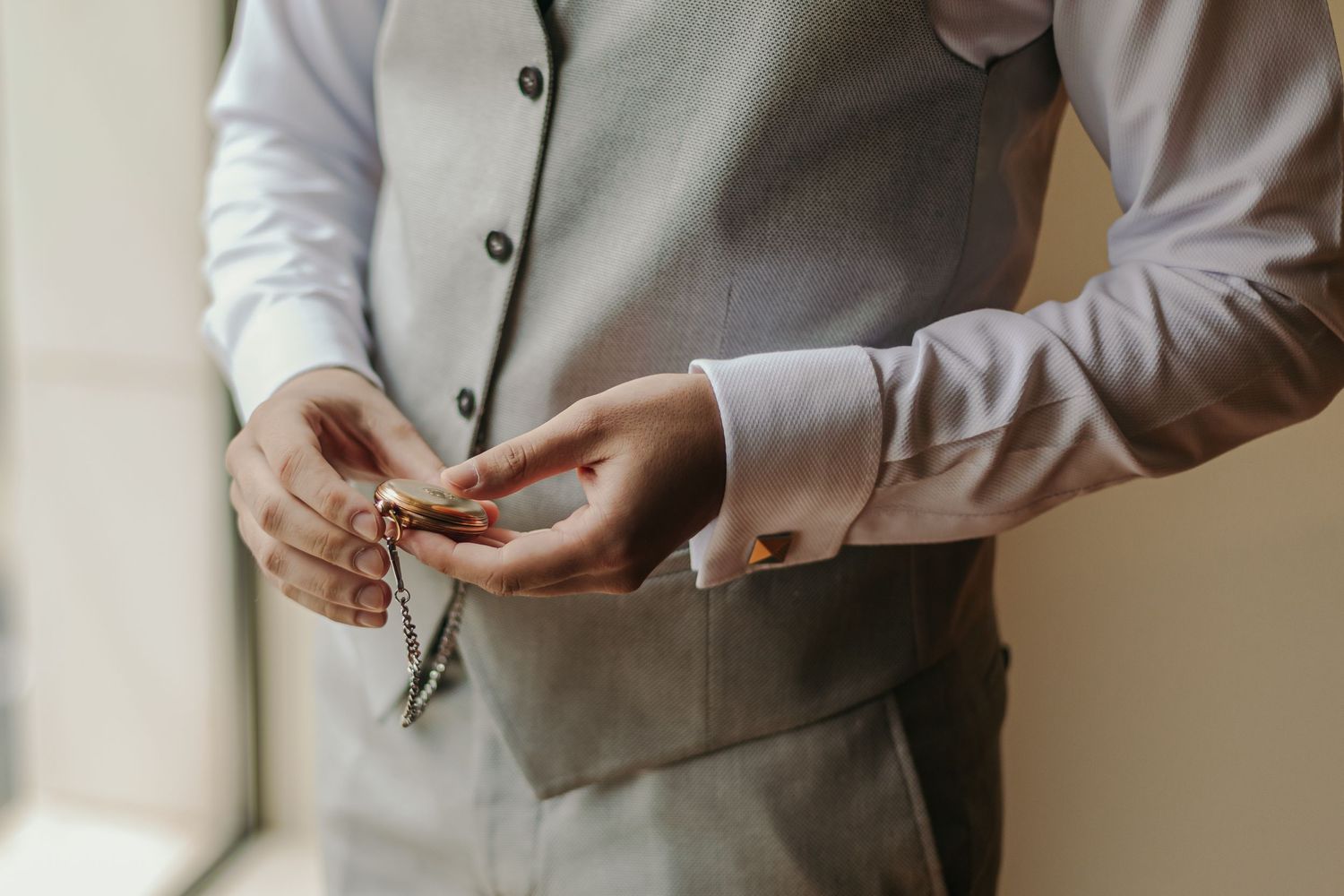 Groom in grey suit vest fastening cufflinks while holding keys before wedding ceremony.