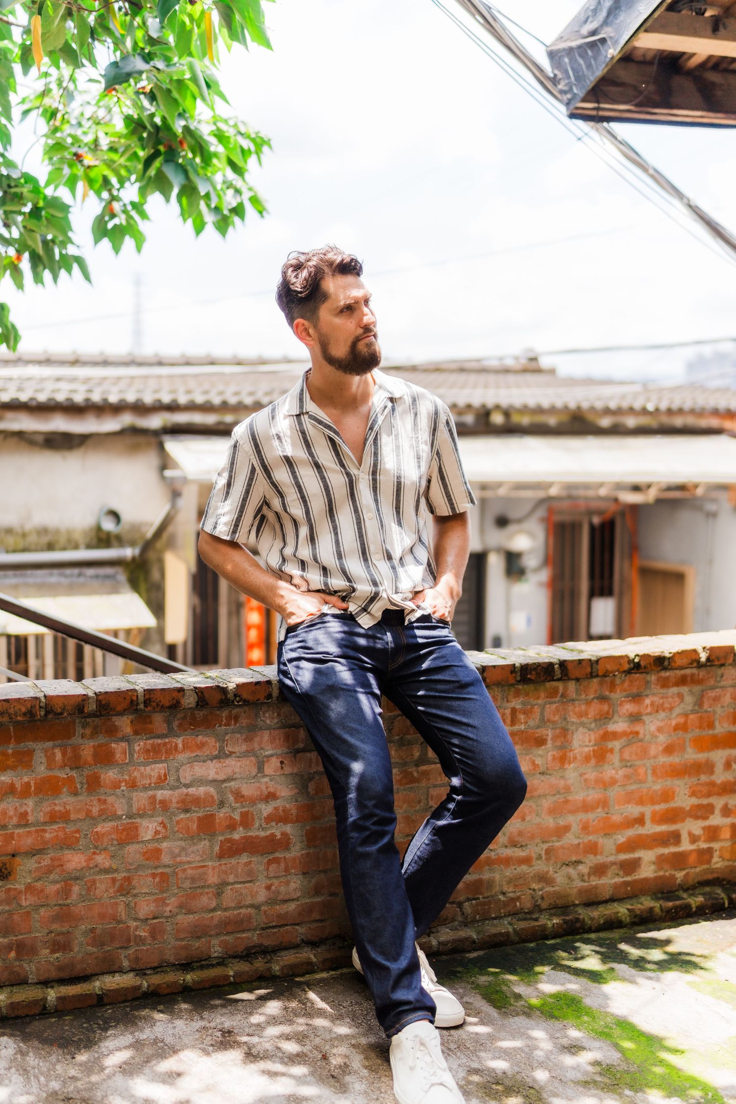 Man in striped shirt sits casually on brick wall looking into distance with greenery overhead.