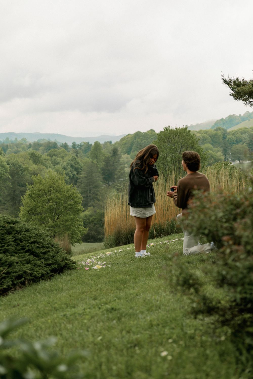 Two people walking on a scenic hilltop path surrounded by green mountains and trees.