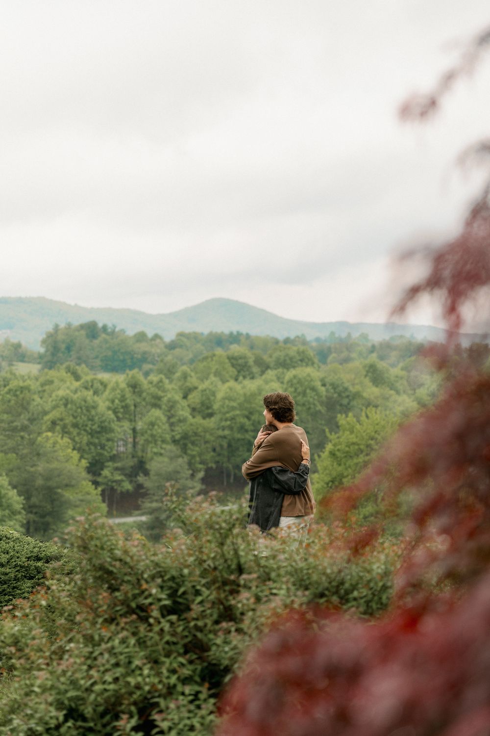 A person sits on a hilltop overlooking a lush green valley and mountains on a cloudy day.