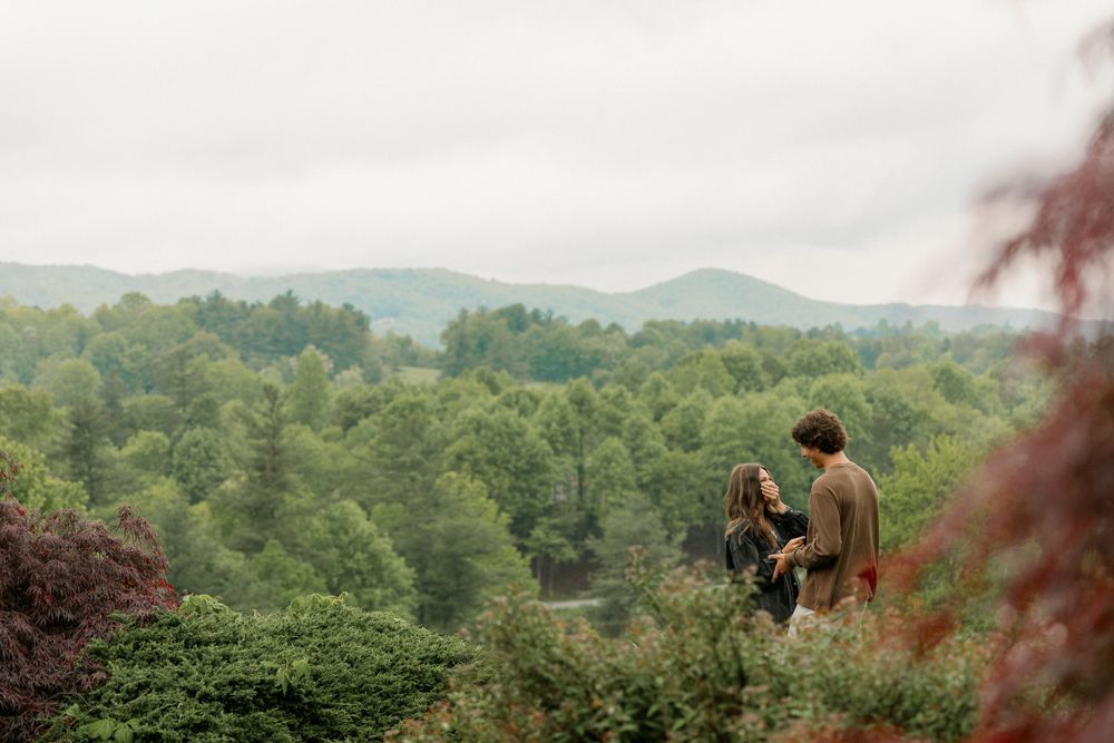 Two people stand together on a scenic overlook with rolling mountains and forests in the distance.