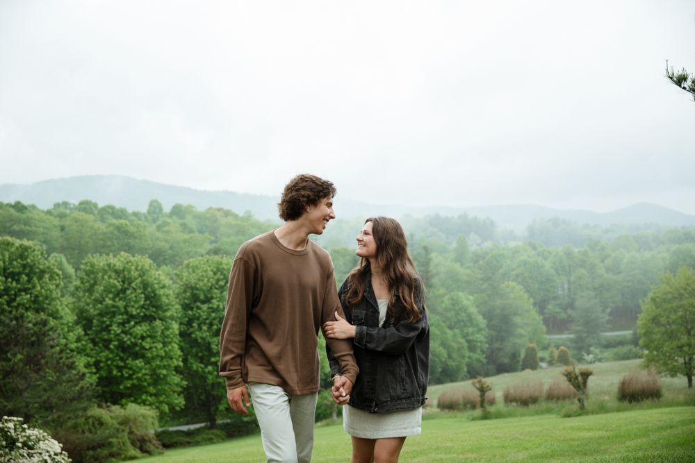 A couple walks hand in hand on a hilltop with a misty mountain landscape behind them.