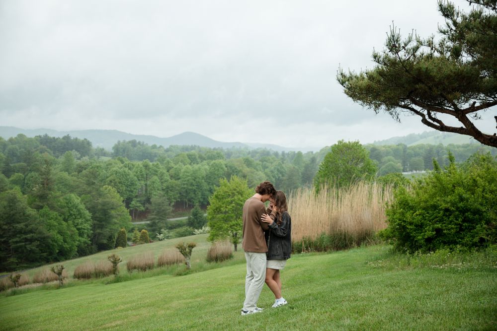 A couple embraces on a grassy hillside overlooking a misty mountain valley.