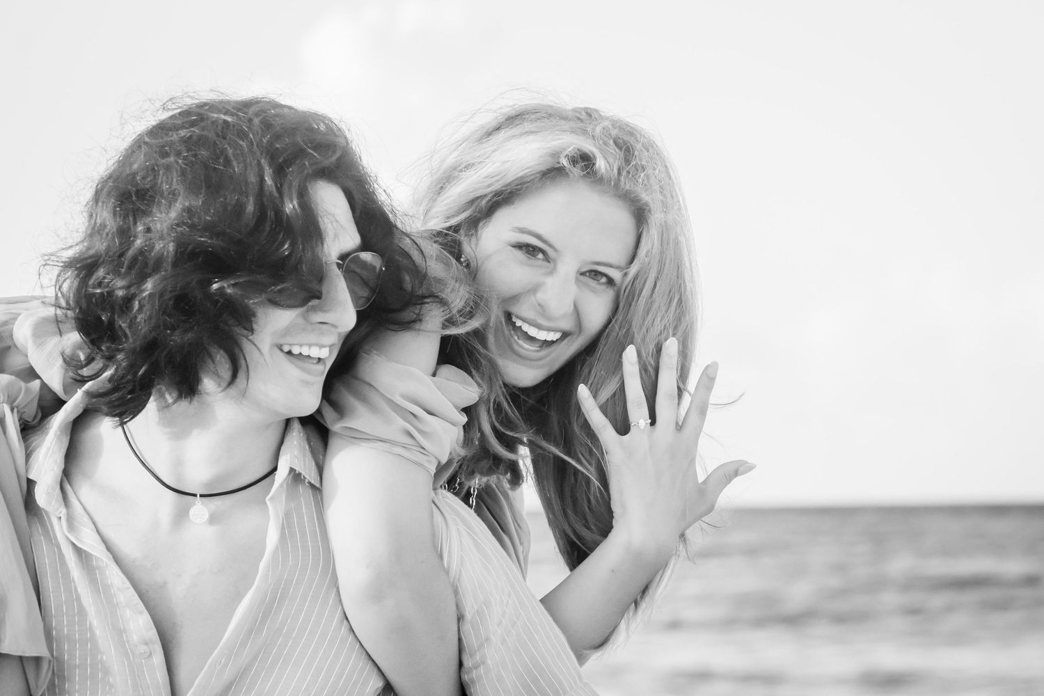 Two women laughing joyfully on a beach, one showing off an engagement ring in black and white photo.