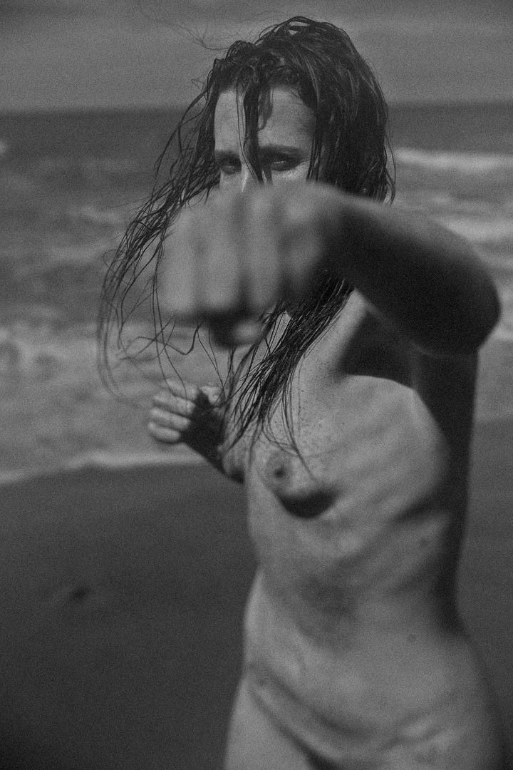Black and white artistic nude photograph of a figure on a beach with wet hair and dramatic shadows.