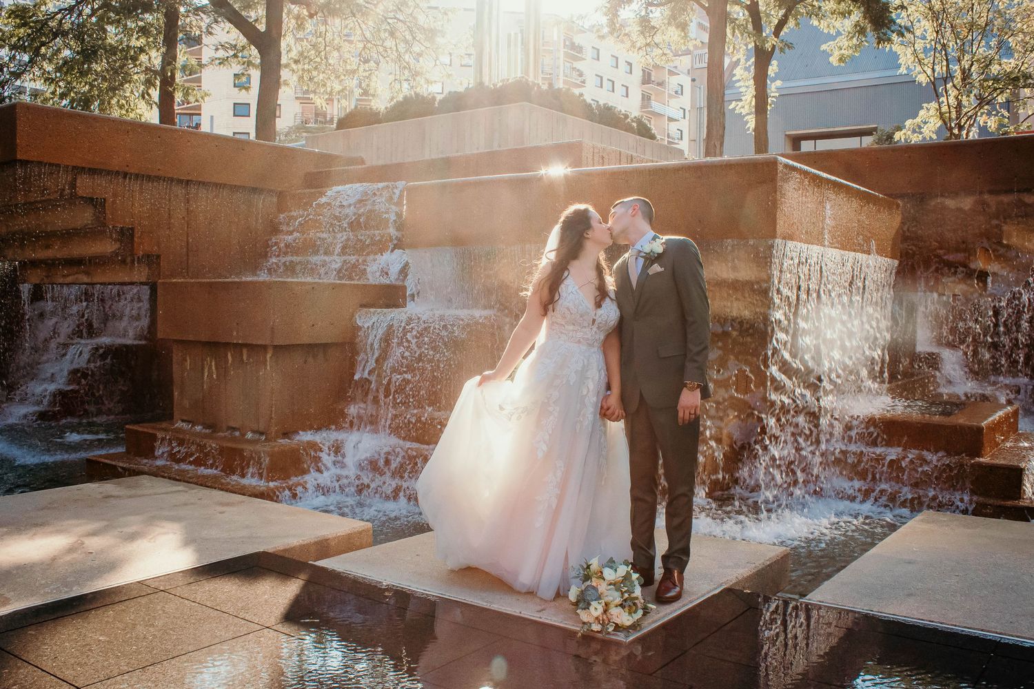 A couple in wedding attire stands in front of a cascading water fountain with sunlight streaming through the mist.