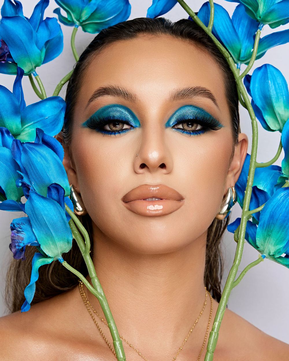 A close-up portrait of a model with bold blue eye makeup, glossy neutral lips, and vibrant blue flowers framing her head