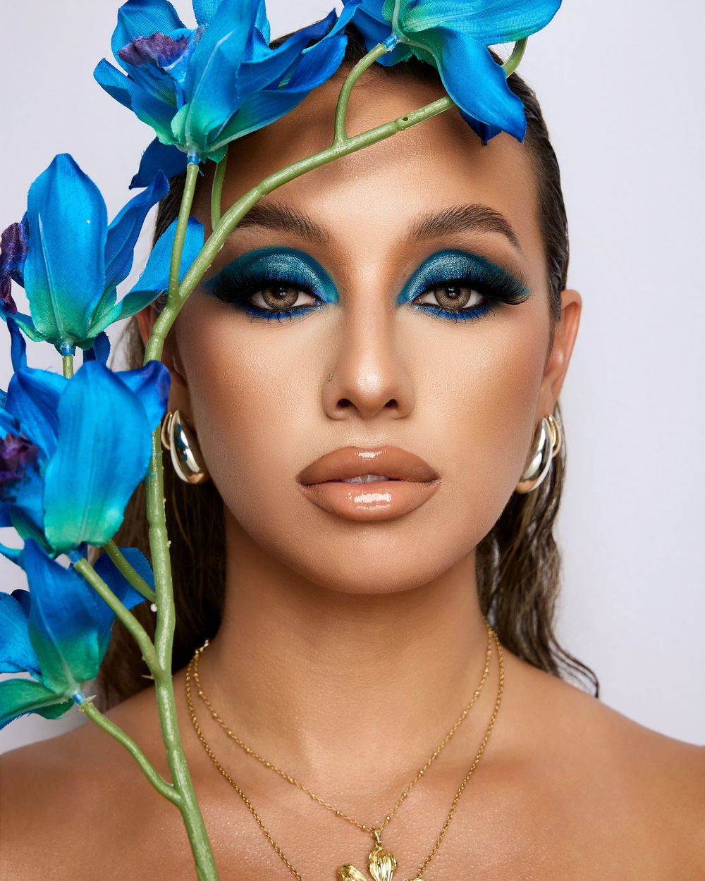A close-up portrait of a model with bold blue eye makeup, glossy neutral lips, and vibrant blue flowers framing her head