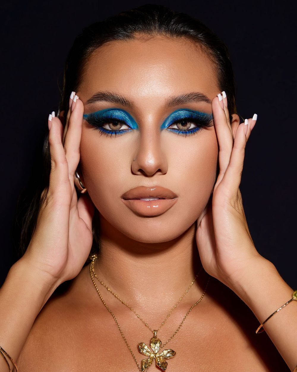 A close-up portrait of a model with bold blue eye makeup, glossy neutral lips, and vibrant blue flowers framing her head