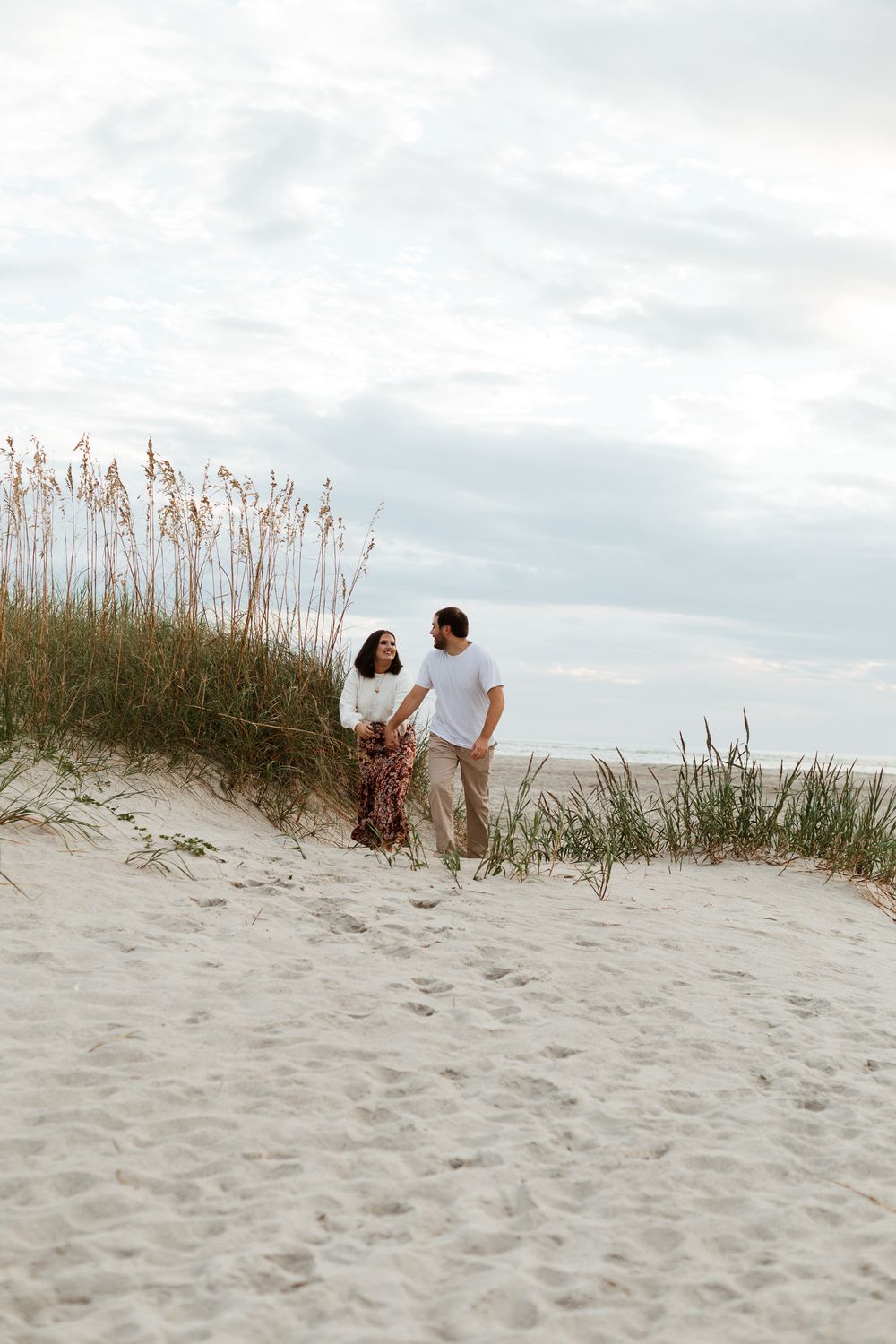 Couple walking together on sandy beach dunes with sea oats during cloudy day.
