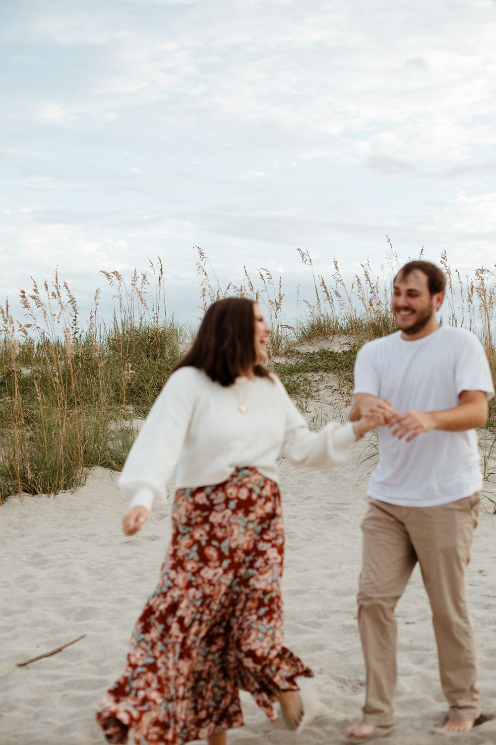 A couple dances playfully on a sandy beach with sea oats swaying in the background at sunset.