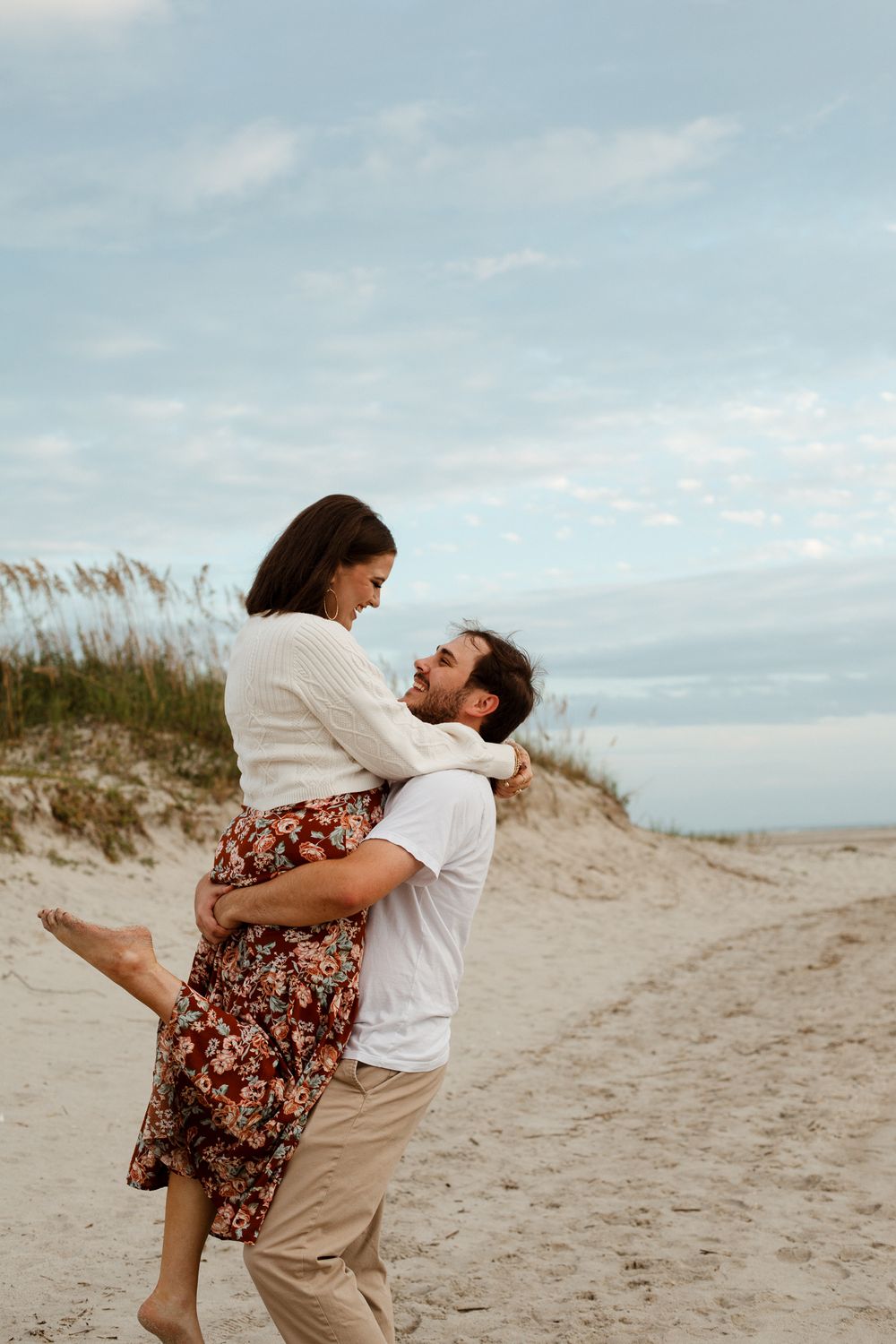 A romantic couple embraces on a beach with sand dunes and coastal vegetation in the background.
