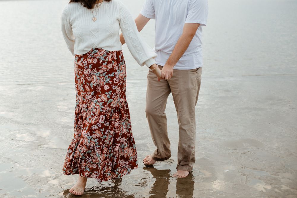 A couple holds hands while standing barefoot on wet sand at the beach.