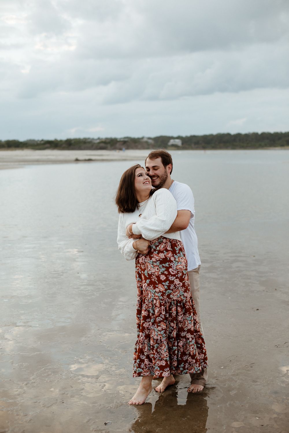 A couple shares an intimate moment on a cloudy beach with water and trees in the background.