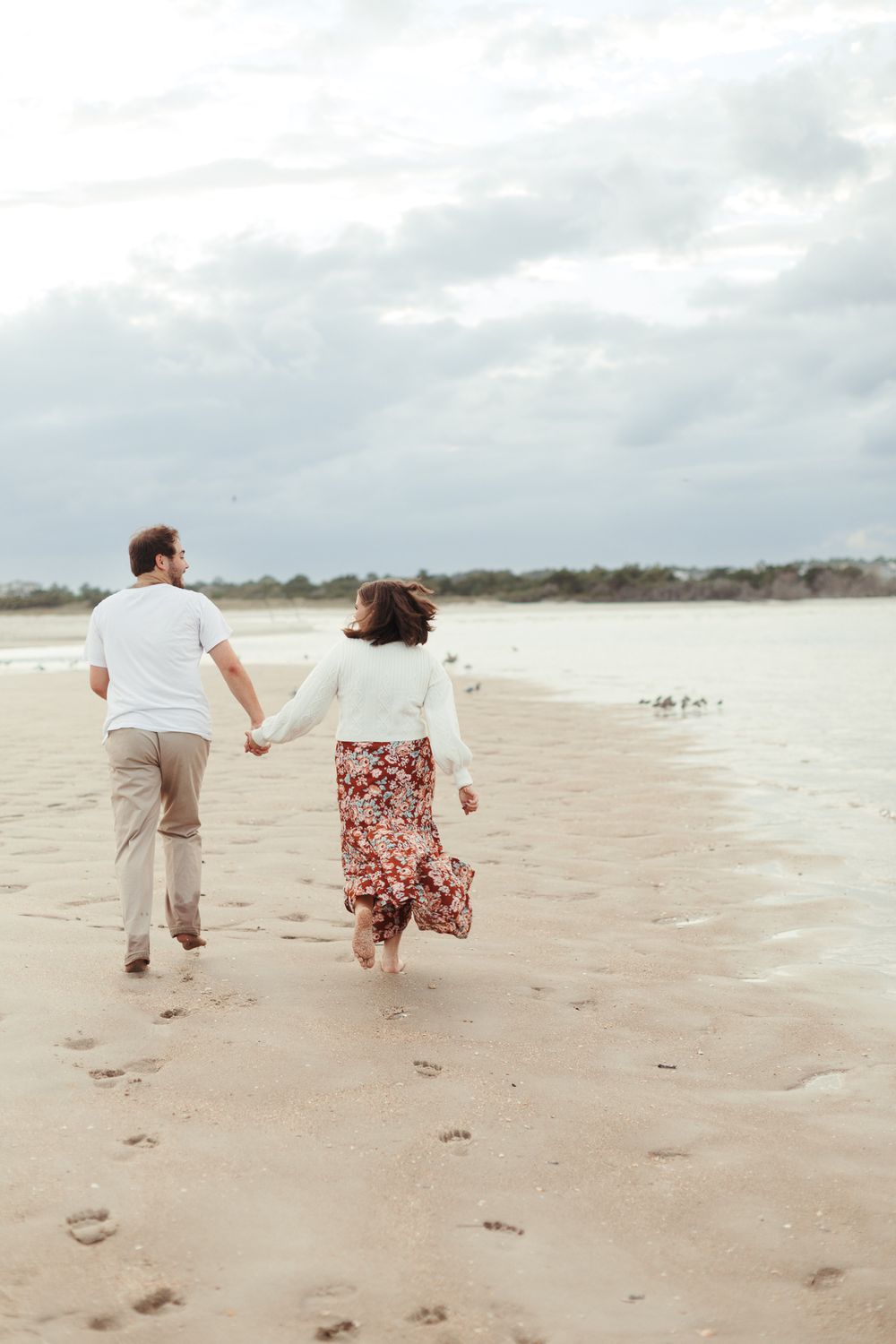A couple runs hand in hand along an empty beach during an overcast day.