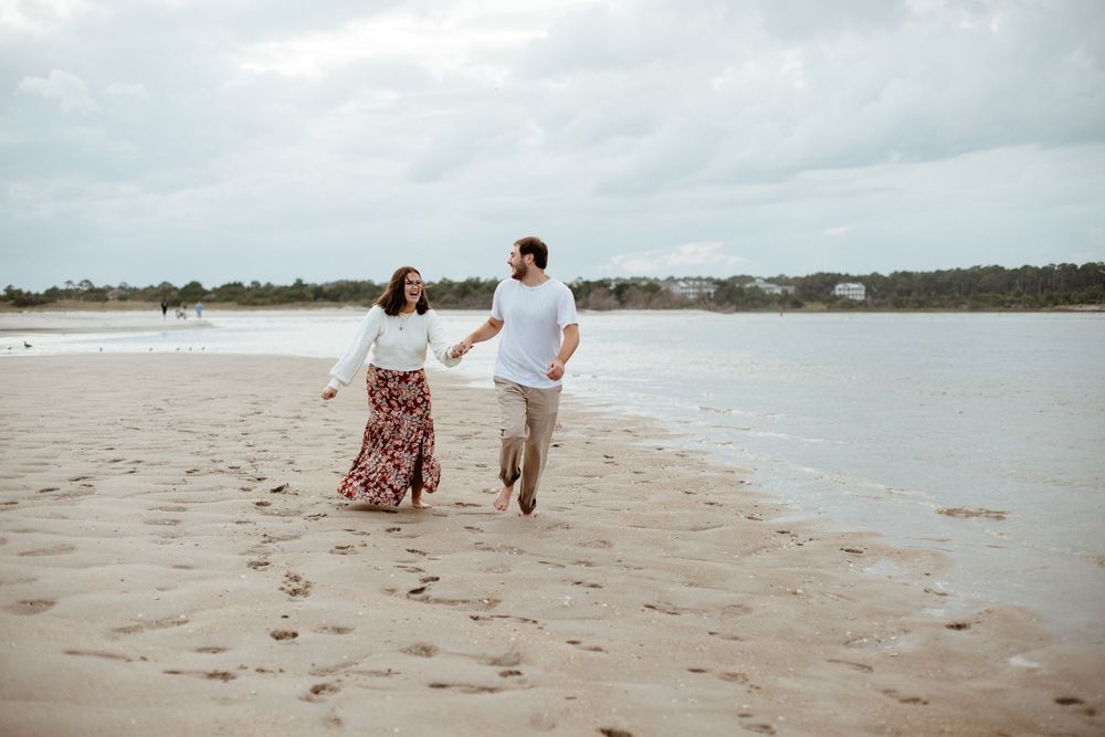 A couple walks together on a sandy beach with coastal homes visible in the distance.