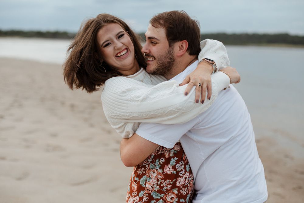 A joyful couple embraces and laughs together on a beach during their photoshoot.
