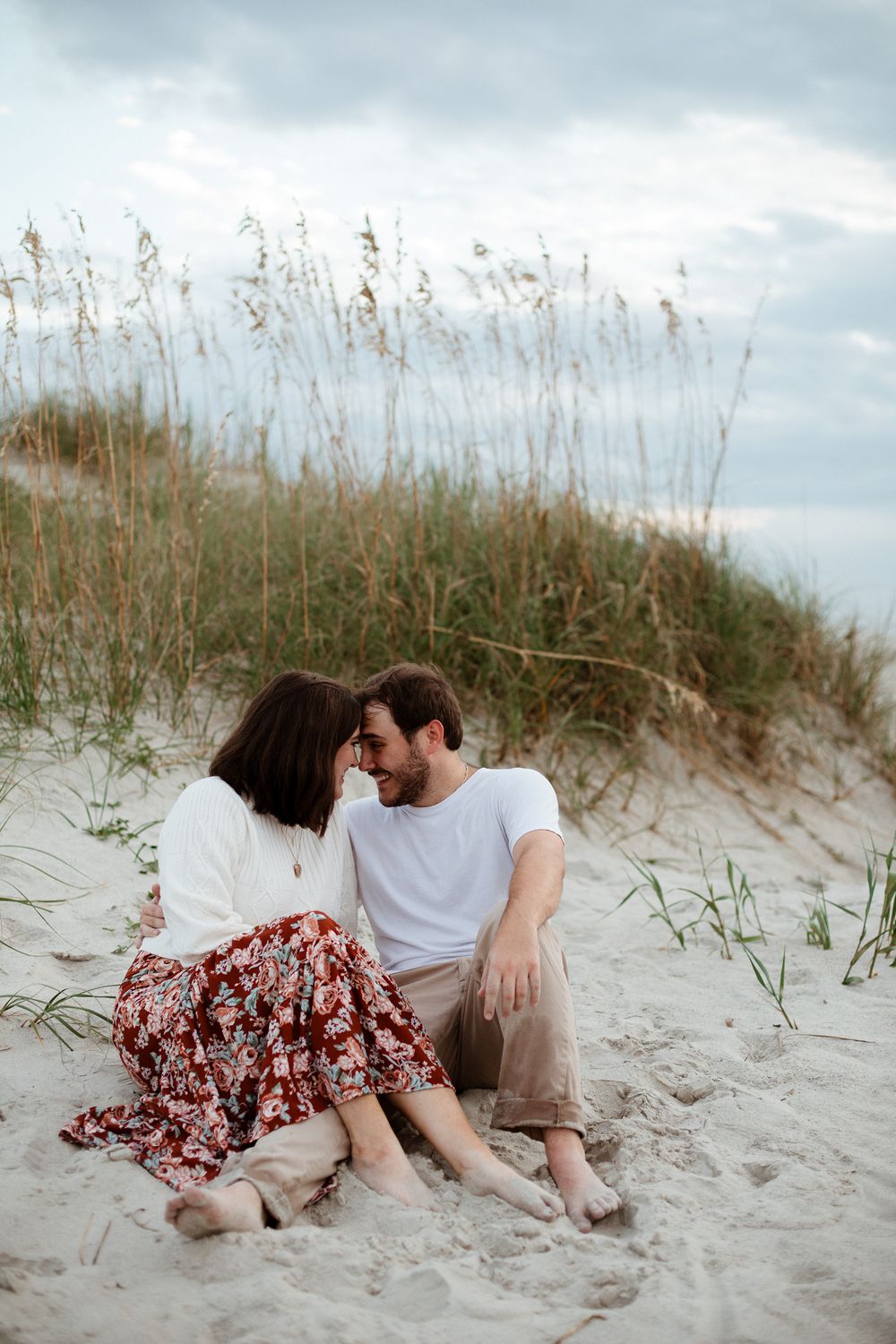 A couple sits intimately on sand dunes surrounded by sea oats during a beach photoshoot.