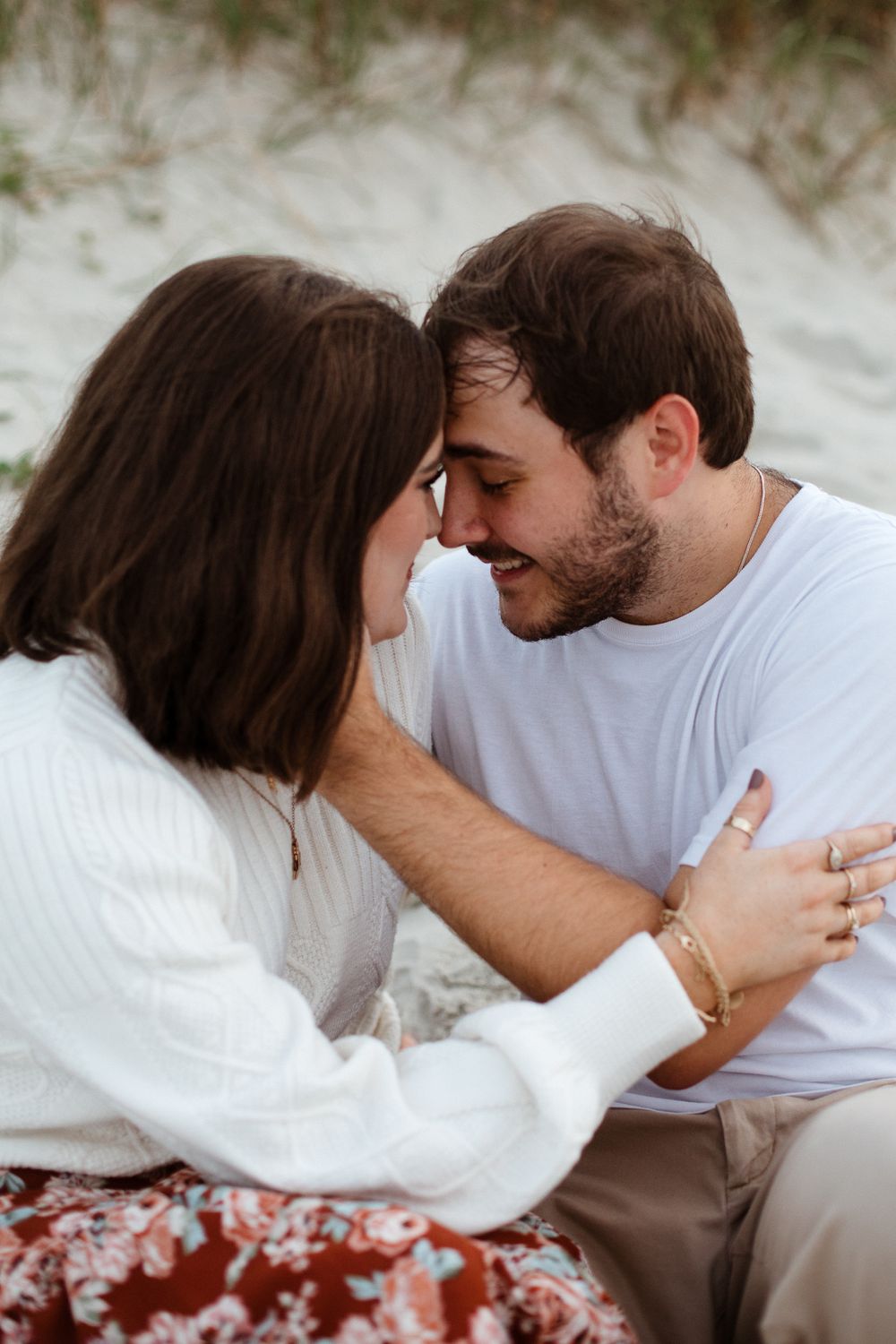 A couple shares a tender moment while sitting on white sand dunes during a beach photoshoot.
