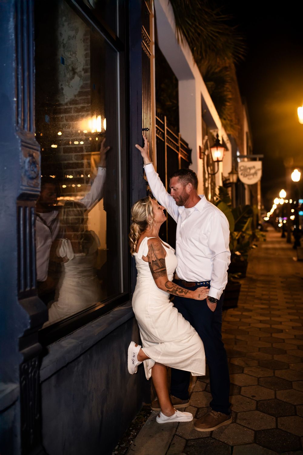 A couple shares a romantic moment outside a storefront at night with glowing street lamps and illuminated windows.