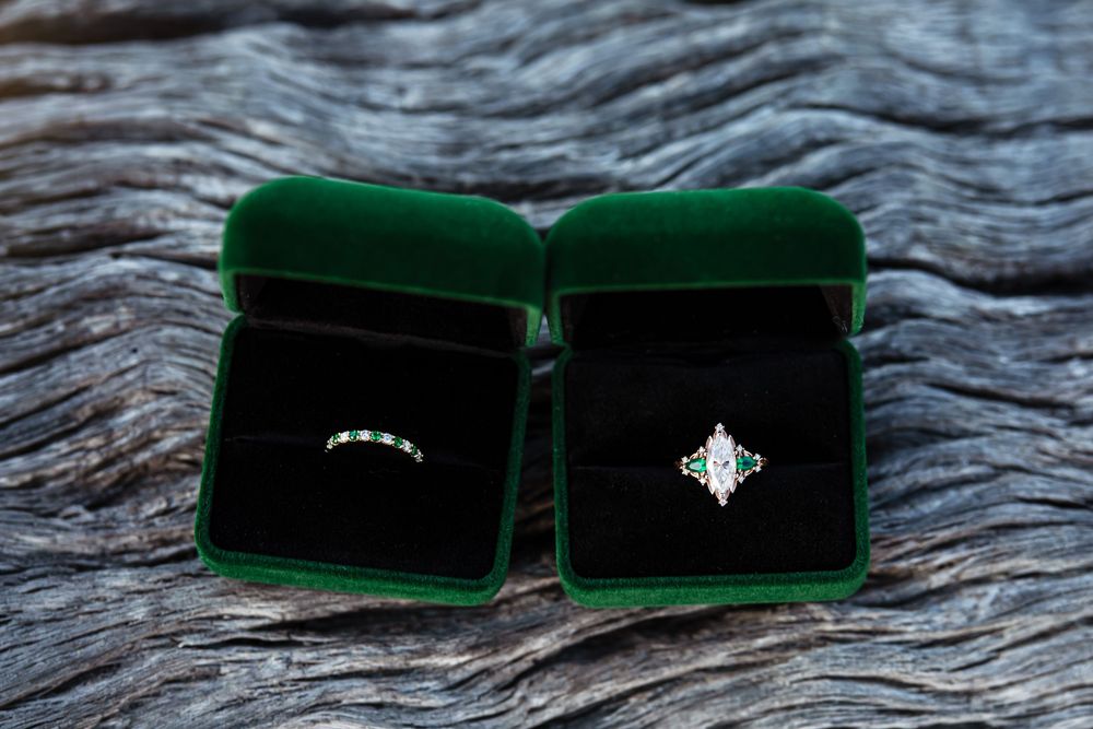 Two elegant rings displayed in open green velvet jewelry boxes on a rustic wood surface, featuring a band and a gemstone ring.