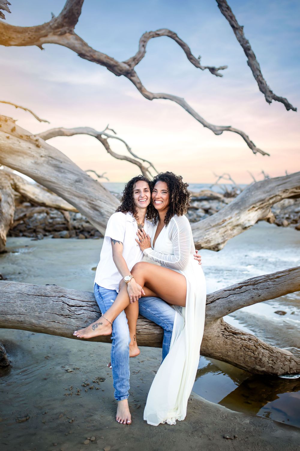 Two women sitting on driftwood at a beach during sunset, smiling and embracing in white tops and jeans.